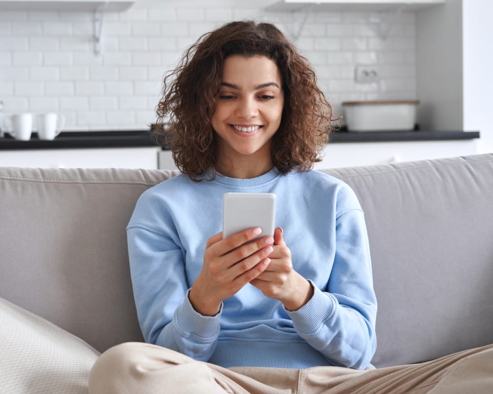A teenage girl sits on the couch and smiles while looking at her smartphone.