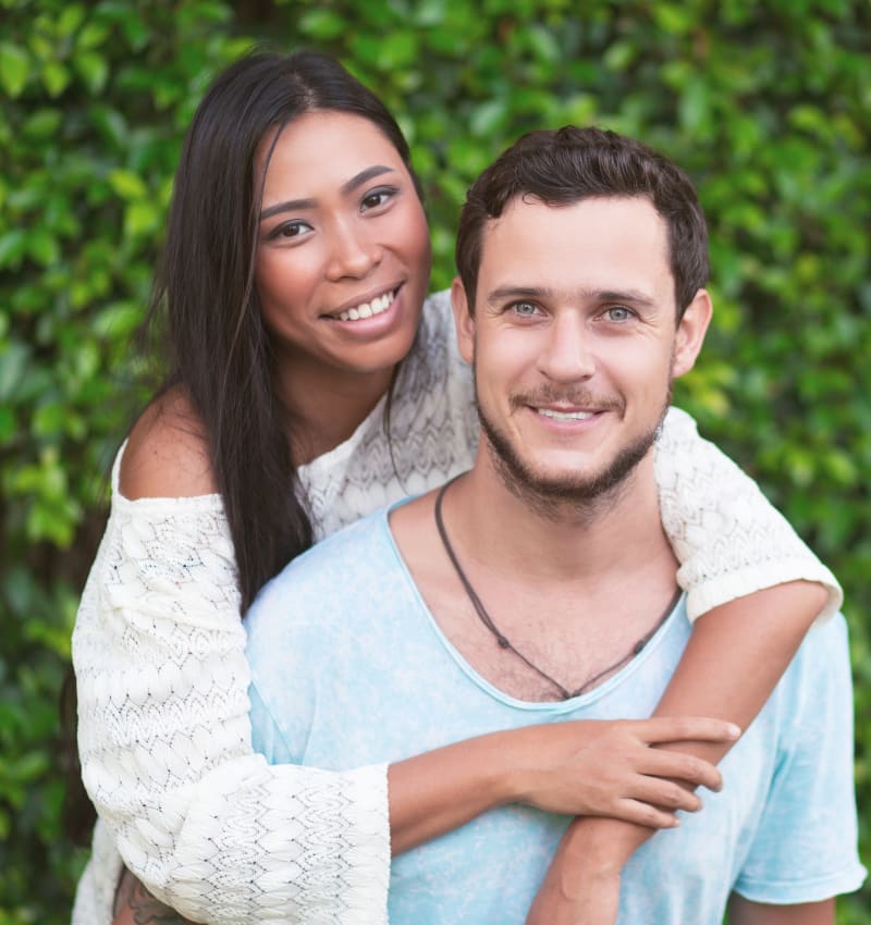 A young adult woman hugs a young adult man from behind. They are both facing the camera and smiling.