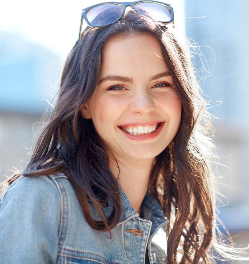 A teenage girl with sunglasses on her head is standing outside and smiling at the camera.
