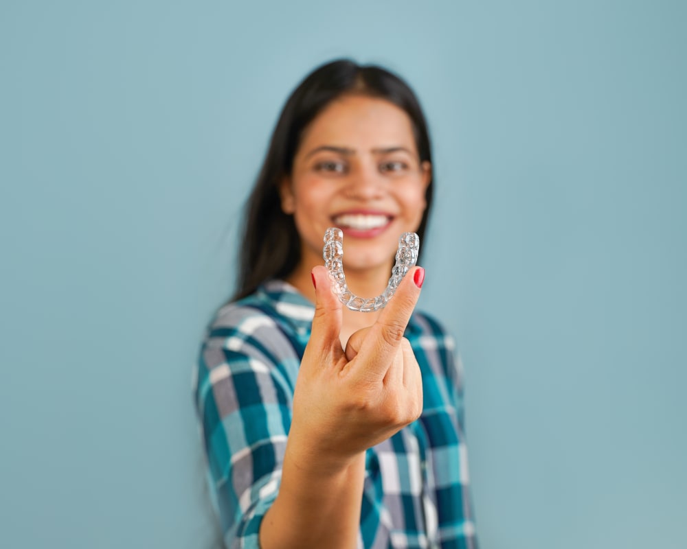 In the background of this photo, a woman is smiling. In the foreground, she is holding an Invisalign clear aligner.