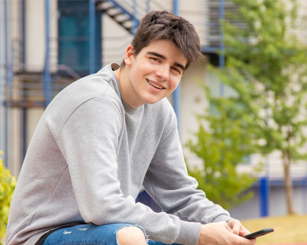 A teenage boy with braces smiles at the camera while using his smartphone.