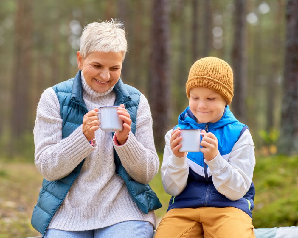 A grandmother and her grandson look into mugs. They are sitting outside in a wooded area wearing warm clothing.