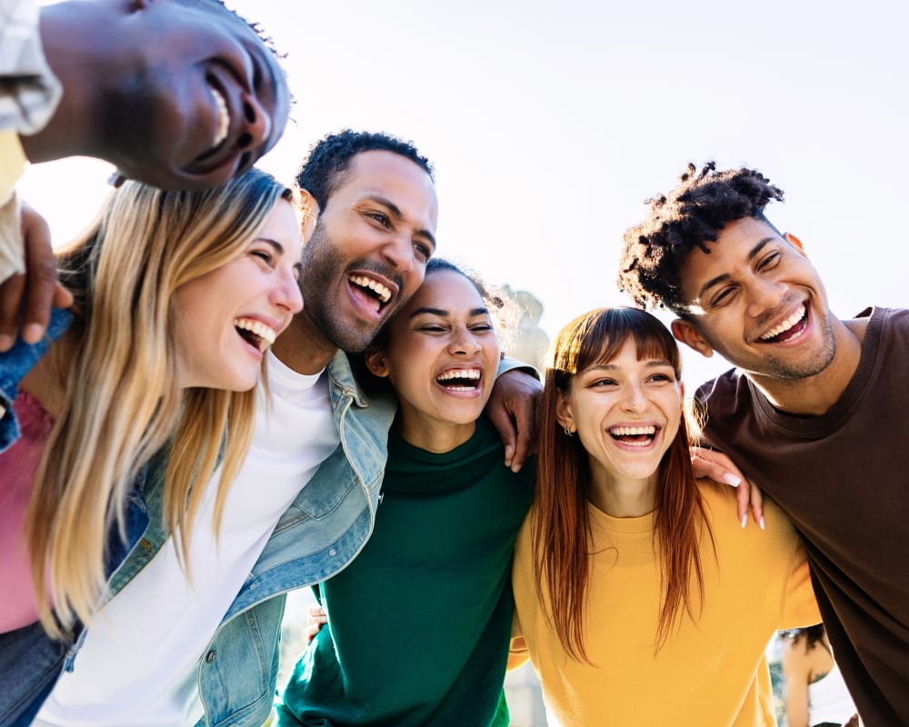 A diverse group of young adults has their arms around each other and is smiling.
